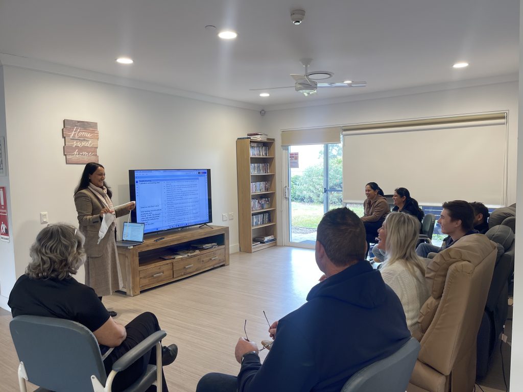 A nurse educator from Uprety Home Care delivers bedside clinical training to a group of support staff in a community care home. The educator is standing near a large screen displaying clinical content, while the attendees, seated in a semi-circle, are engaged and smiling. The setting is a bright, home-like room with a “Home sweet home” sign on the wall and natural light coming through a window.